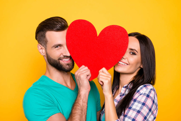 Portrait of lovely cheerful couple looking out big carton paper heart figure having beaming smiles isolated on bright yellow background. Fall in love true feelings concept - Photo, image