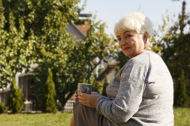 A beautiful blonde pensioner woman sitting and smiling in the garden with a cup of tea and cookies. Copy space. - Foto, Bild