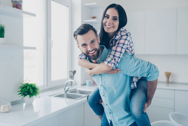 Portrait of cheerful joyful couple, handsome man carrying on back pretty woman standing in modern white kitchen looking at camera enjoying tome together - Photo, image