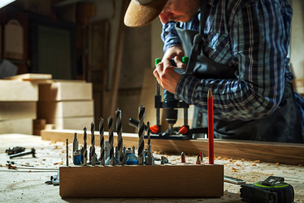 Experienced carpenter in work clothes and small buiness owner  is carving a wooden board on an  modern  hand drill in a light workshop side view, in the background a lot of tools - Photo, Image