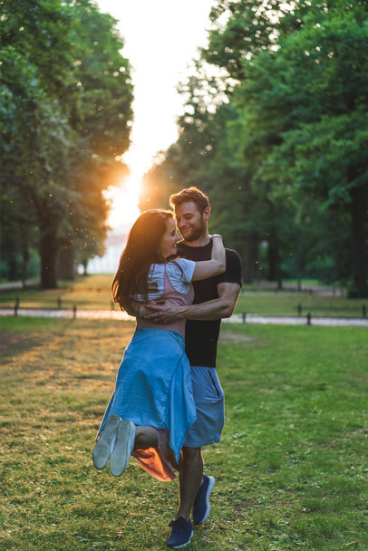 sonriente hombre sosteniendo joven novia en parque durante la puesta del sol
 - Foto, Imagen