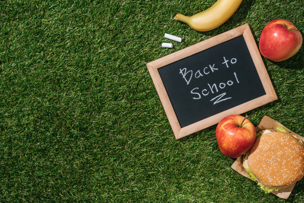 flat lay with blackboard with back to school lettering, pieces of chalk, banana, apples and burger on green grass - Photo, Image