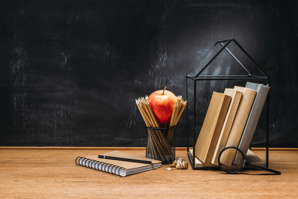 close up view of fresh apple, magnifying glass, notebook and books on wooden surface with empty blackboard behind - Foto, afbeelding