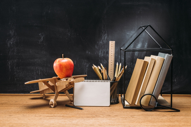 close up view of apple on wooden toy plane, notebook, books and pencils on tabletop with empty blackboard behind - Photo, Image