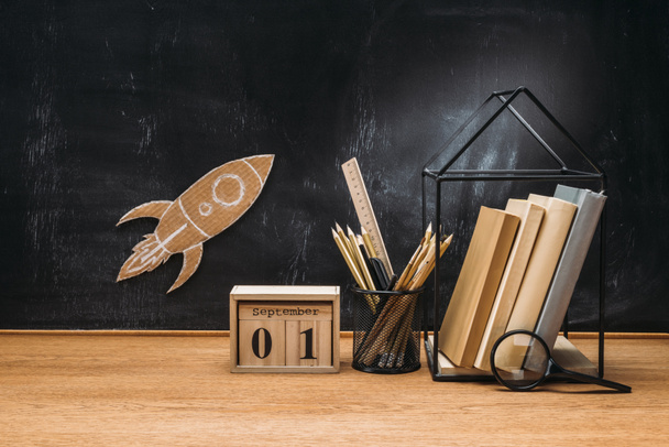 close up view of cardboard rocket on blackboard, calendar, magnifying glass and books on wooden tabletop - Zdjęcie, obraz