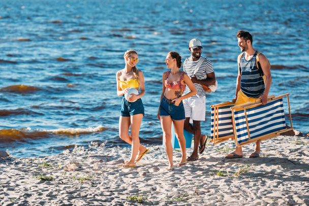 улыбающиеся молодые многонациональные друзья с пляжными принадлежностями прогуливаясь по песчаному побережью моря
  - Фото, изображение