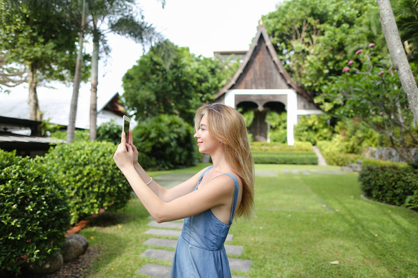 Mujer joven tomando fotos por teléfono inteligente y de pie con la casa del complejo en el fondo
. - Foto, imagen
