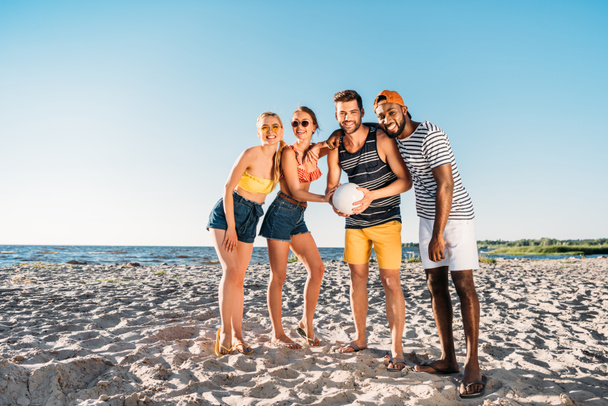 happy young multiethnic friends holding ball and smiling at camera on beach  - Fotografie, Obrázek