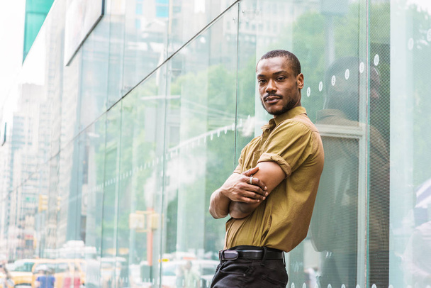 Young African American Man with beard, wearing green short sleeve shirt, black pants, arms crossing in front, standing against glass wall with reflections on street in Manhattan, New York, relaxing - Photo, Image