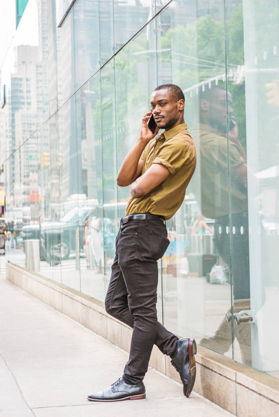 Young African American Man with beard, wearing green short sleeve shirt, black pants, leather shoes, standing against glass wall with reflections on street in New York, relaxing, talking on cell phone - Foto, Imagen