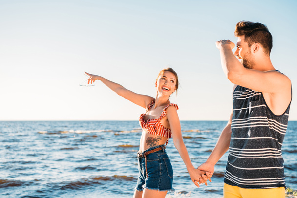 happy young couple holding hands and looking away while walking together on beach - Foto, Imagen
