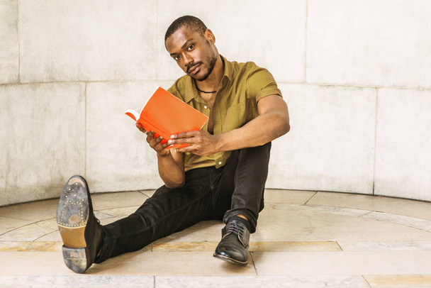 junger afrikanisch-amerikanischer Mann mit Bart studiert in New York, trägt grünes Kurzarmshirt, schwarze Hose, Lederschuhe, sitzt auf Marmorboden auf dem Campus, liest rotes Buch, schaut weg, denkt nach - Foto, Bild