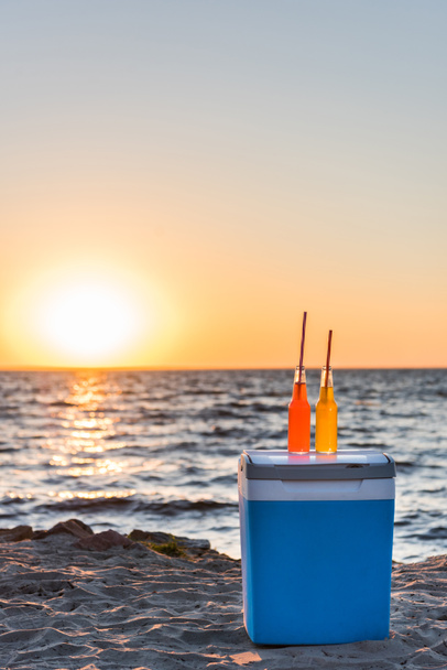 glass bottles with summer beverages and straws on cooler at sandy beach at sunset - Фото, зображення