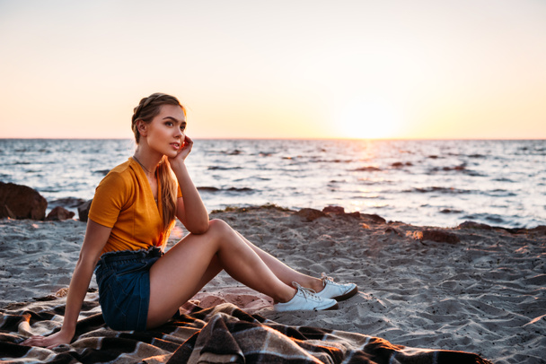 pensive young woman sitting on plaid at beautiful sea coast at sunset  - Foto, imagen