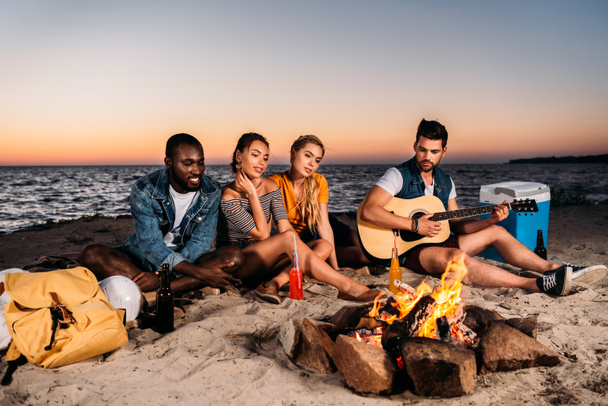 jóvenes amigos multiétnicos disfrutando de la guitarra y pasar tiempo juntos en la playa de arena al atardecer
  - Foto, Imagen