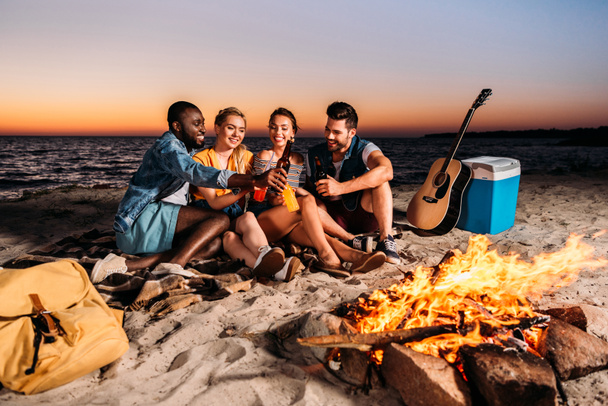 happy multiethnic friends clinking glass bottles with drinks while spending time together on sandy beach at sunset  - Photo, Image