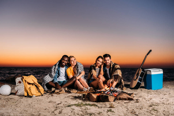 glückliche junge Leute lächeln in die Kamera, während sie bei Sonnenuntergang zusammen am Sandstrand sitzen  - Foto, Bild