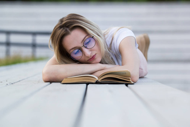 Όμορφη νεαρή γυναίκα διαβάζοντας ένα βιβλίο που λέει ψέματα στον πάγκο στο πάρκο - Φωτογραφία, εικόνα