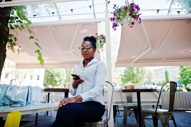 Opotřebení africká americká dívka v brýlích s mobilním telefonem u venkovní caffe. - Fotografie, Obrázek
