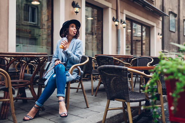 Beautiful young woman having orange juice in outdoor cafe and showing tongue to passer-by. Stylish fashionable outfit - Foto, Imagen
