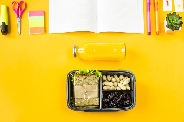 Lunchbox mit Essen - ein Sandwich, Nüsse und Beeren - steht neben dem Notizbuch, Stiften und anderen Utensilien für das Studium. Mahlzeiten zwischen den Unterrichtsstunden. Draufsicht, flache Lage - Foto, Bild