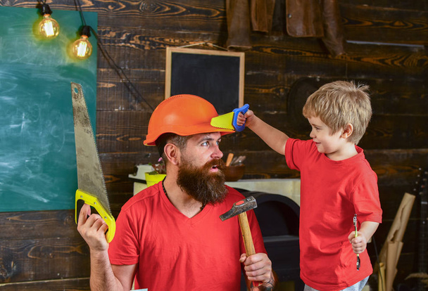 Father, parent with beard in protective helmet teaching little son to use different tools in school workshop. Boy, child cheerful playing with toy saw, learning use tools with dad. Fatherhood concept - Photo, Image