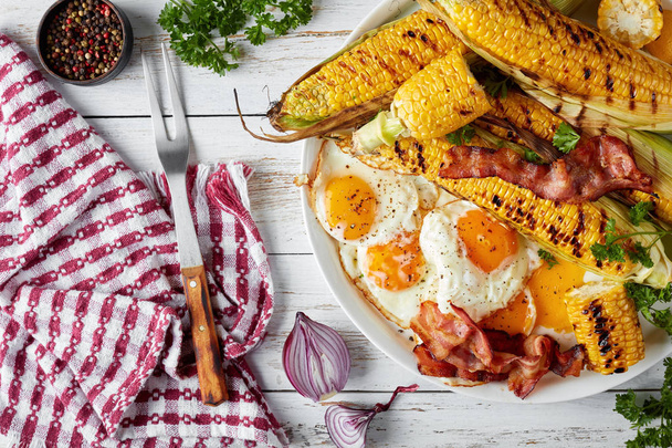 grilled corn in cobs with fried eggs and bacon slices on a white dish on an old rustic table with napkin and fork, horizontal view from above, flat lay - Photo, Image