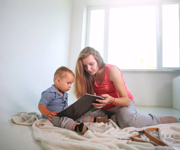 Mother with a little baby son in a room reading a book at home - Фото, изображение