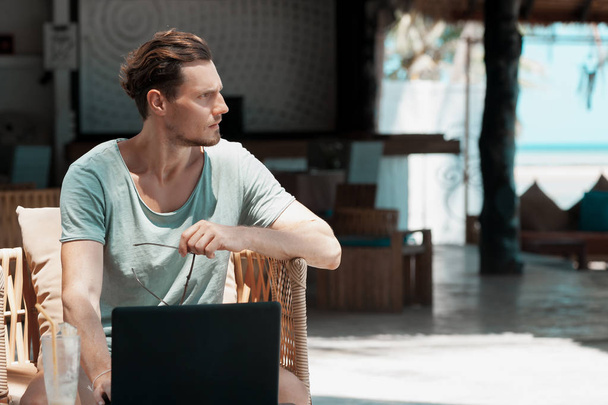 Человек фрилансер с ноутбуком и мобильным телефоном сидит в кафе на открытом воздухе
. - Фото, изображение