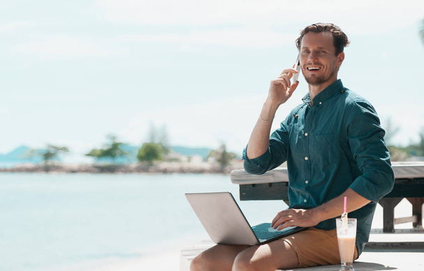 Homem freelancer com laptop e telefone celular sentado na praia
 - Foto, Imagem
