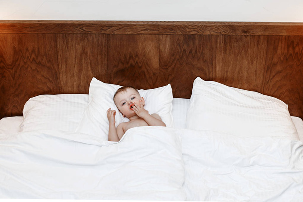 L'enfant est au lit. Garçon sur un grand lit entre une couverture et un oreiller. Un petit bébé est assis au lit et mange. L'enfant qui se livre
. - Photo, image