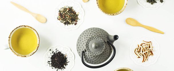 Gray iron kettle among different kinds of dry tea and cups with a ready drink on a white table. Top view, flat lay - Fotó, kép