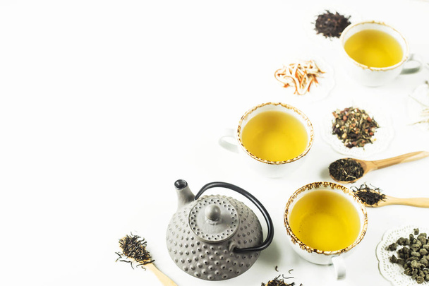 Gray iron kettle among different kinds of dry tea and cups with a ready drink on a white table. Top view, flat lay. Copy space - Photo, Image