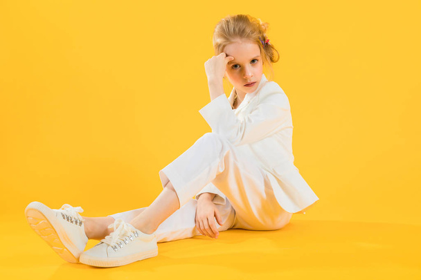 Дівчина-підліток у білому одязі сидить, розтягуючи ноги вперед на жовтому тлі
. - Фото, зображення