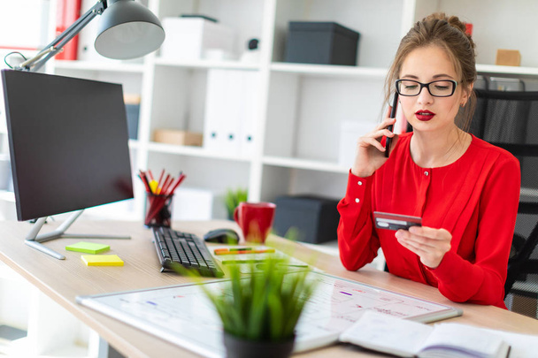 Una joven está sentada en el escritorio de la oficina, con una tarjeta bancaria y un teléfono en la mano.
. - Foto, imagen