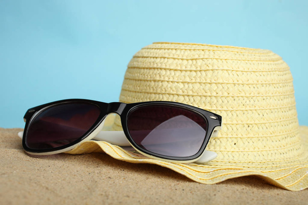 Naarashattu ja aurinkolasit makaavat hiekalla sinisellä pohjalla
. - Valokuva, kuva