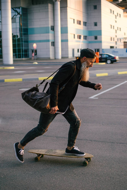 Stylish handsome middle-aged man with long gray beard riding on longboard during sunset  on urban background - Photo, Image