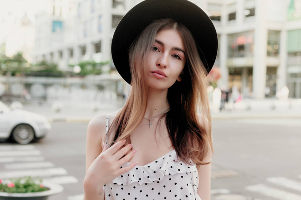 young teenage girl in a white dress and hat. style of the big city. Sunset sky and rays of the sun. Natural beauty pretty woman in black fedora hipster hat, on warm summer evening sunset in big city - Fotó, kép