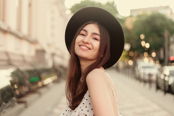 young teenage girl in a white dress and hat. style of the big city. Sunset sky and rays of the sun. Natural beauty pretty woman in black fedora hipster hat, on warm summer evening sunset in big city - Φωτογραφία, εικόνα