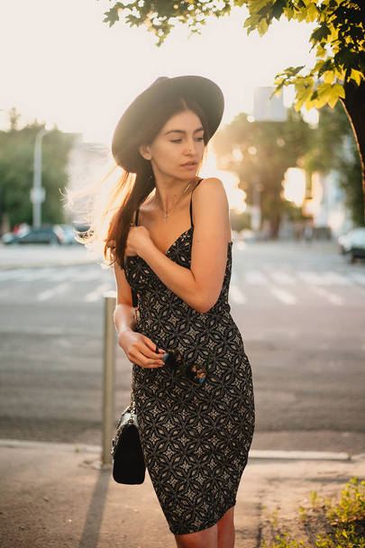 young teenage girl in a white dress and hat. style of the big city. Sunset sky and rays of the sun. Natural beauty pretty woman in black fedora hipster hat, on warm summer evening sunset in big city - Fotó, kép