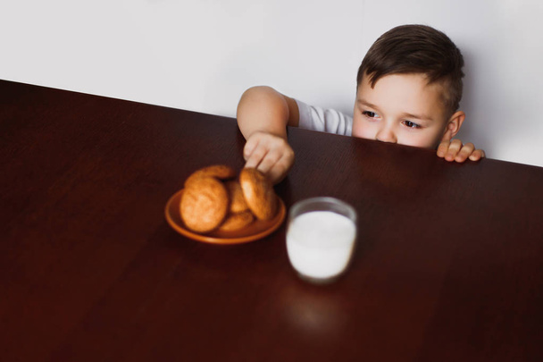 A criança tenta roubar biscoitos de aveia e leite caseiro da mesa na cozinha. Um café da manhã saudável
. - Foto, Imagem
