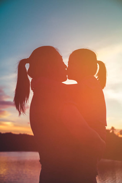 Silhouette of mother carrying and kissing her daughter. Happy family enjoying view in the morning with sunlight, cozy mood. Colorful pastel sky sunrise, serenity background. Travel on vacation. - Foto, Imagen