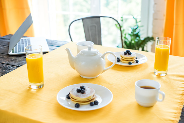 Nahaufnahme von Pfannkuchen, Tassen Tee und Gläsern Saft zum Frühstück auf dem Tisch mit gelber Tischdecke - Foto, Bild
