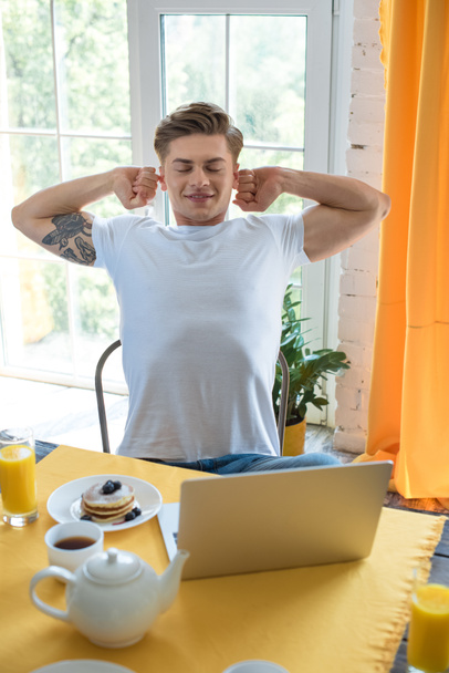 retrato de un joven que se extiende mientras está sentado en la mesa con el desayuno y el ordenador portátil en casa
 - Foto, Imagen