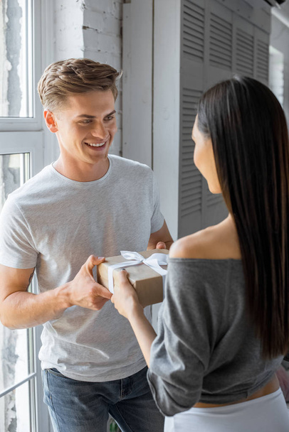 sorrindo caucasiano homem apresentando embrulhado presente para ásia namorada em casa
 - Foto, Imagem