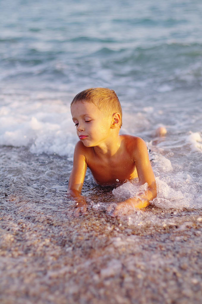 Ein fröhlicher Junge spielt in den Wellen am Strand. fröhlicher Junge badet bei Sonnenuntergang in den Wellen des Meeres - Foto, Bild