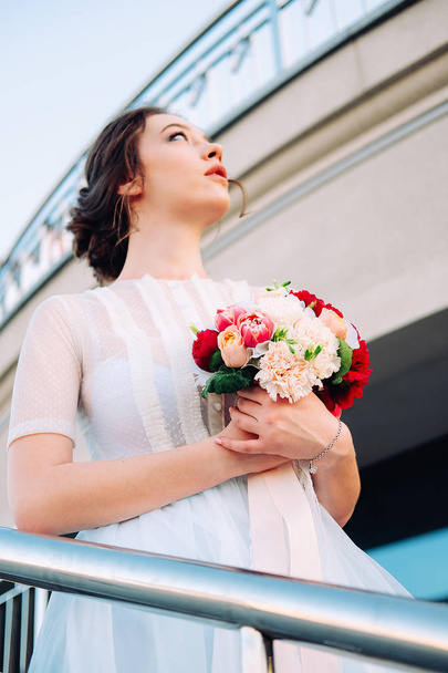 Adorable young bride with bridal bouquet made of peonies, roses, freesia flowers and wearing white tulle wedding dress  standing on the staircase - Фото, изображение