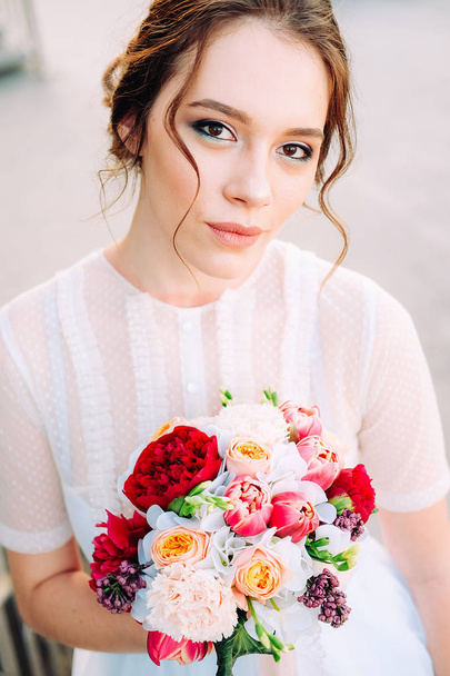 Adorable young bride with bridal bouquet made of peonies, roses, freesia flowers and wearing white tulle wedding dress  standing on the staircase - Foto, Imagem