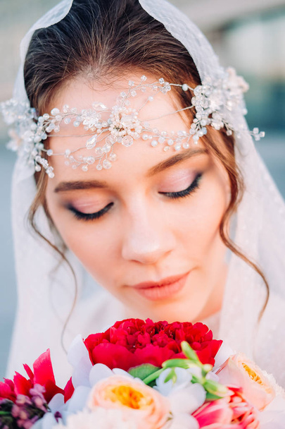 wunderschöne Braut im östlichen Stil mit weißem Schleier und Haarteil, die Brautstrauß hält und bei Sonnenuntergang im Freien posiert - Foto, Bild