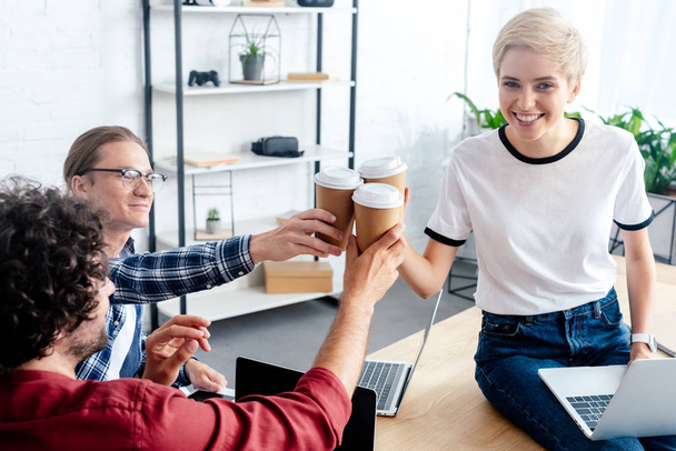 улыбающиеся молодые коллеги звонят бумажными стаканчиками во время использования ноутбуков в офисе
 - Фото, изображение